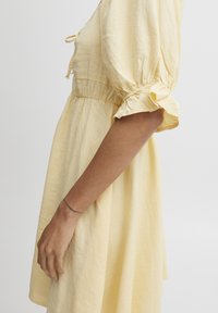 Rochie din in galben, cu manşete bufante, talie strânsă şi detaliu cu şnur la gât. Textură netedă, croială până la genunchi şi culoare delicată.