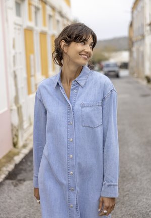Femme en robe en jean bleu clair debout dans une rue calme, regardant vers sa gauche avec un léger sourire.