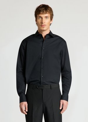 Giovane uomo con capelli castano chiaro che indossa una camicia nera a maniche lunghe con bottoni e pantaloni neri, in piedi davanti a uno sfondo chiaro e uniforme.