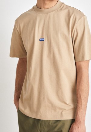 Beige Baumwoll-T-Shirt mit rundem Ausschnitt und kurzen Ärmeln, versehen mit einem blauen "HUGO"-Logo auf der Brust. Glatte Textur, lockere Passform.