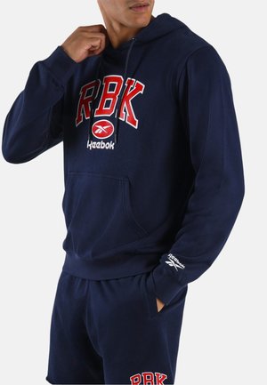 Mann trägt navyblauen Reebok-Hoodie und passende Shorts, mit rotem und weißem RBK-Logo auf Brust und Shorts, Hand passt den Hoodie an.