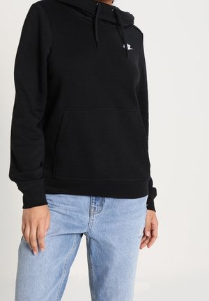Personne portant un sweat à capuche noir avec une poche avant et un jean bleu clair, debout devant un fond clair uni.