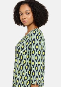 Grüner Tunika mit marineblauem und gelbem geometrischem Muster; verfügt über dreiviertellange Ärmel und einen Rundhalsausschnitt; glatte Stofftextur.