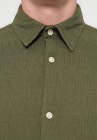 Tuvplāns cilvēkam, kurš valkā olīvu zaļu kreklu ar faktūru, kas aizpogājams ar pogām, ar punktētu apkakli un trim redzamām baltām pogām.