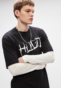 Μαύρη μπλούζα T-shirt με άσπρο γραφικό κείμενο "HUGO," επάνω από άσπρη μακρυμάνικη μπλούζα. Μοντέλο που φορά ασημένια αλυσίδα στο λαιμό.