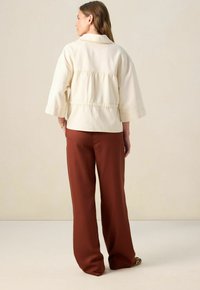 Vrouw die met haar rug naar de camera staat, gekleed in een losse crèmekleurige jas met wijde mouwen en roestkleurige wijde broek.