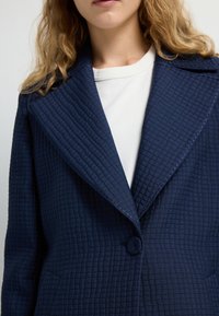 Blazer trapuntato navy con ampio colletto, tessuto testurizzato, chiusura a bottone singolo e tasche laterali. Sotto indossa una maglietta bianca a collo tondo.
