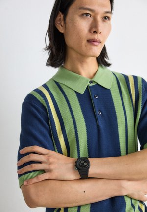 Polo a maniche corte blu navy, verde e giallo a righe; presenta un colletto verde e una chiusura con tre bottoni. Orologio nero con quadrante rotondo.
