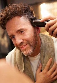 Een man met krullend haar en een kort geschoren baard houdt haartrimmers in zijn hand terwijl zijn haar wordt geknipt. Hij draagt een lichte shirt en een handdoek.