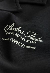 Tissu noir avec texte blanc « Studios Italia ESTD. MCMLXXIII MEMBERS ORIGINALS RESERVE » imprimé dans différentes polices.