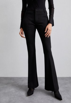 Osoba ubrana w dopasowane czarne spodnie z wysokim stanem i rozszerzanymi nogawkami, przejrzystą czarną bluzkę z długim rękawem oraz czarne szpilki z ostrym czubkiem, stojąca na szarej podłodze.