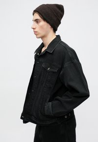 Casaco de ganga preto com um corte relaxado, grande colarinho, bolsos frontais e fechos de botão. Usado com um gorro de malha castanho escuro.