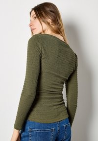 Olive green, long-sleeve top with a textured, gathered design and a wide neckline. Paired with blue denim jeans.
