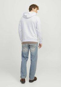 Sudadera de algodón gris con capucha ajustable y puños de canalé, llevada sobre una camisa marrón clara, combinada con jeans azules desgastados y zapatos marrones.