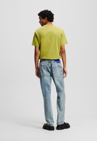 Camiseta verde de manga corta con cuello redondo, combinada con unos vaqueros de mezclilla azul claro que tienen bolsillos traseros y una etiqueta azul en la cintura.