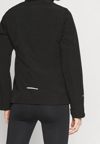 Person iført en sort jakke og sorte leggings, vist fra ryggen, med en lille hvid reflekterende streg på jakken nederst på ryggen.