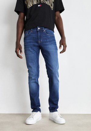 Man draagt slim-fit blauwe spijkerbroek, witte sneakers en een zwart T-shirt met de tekst "Tommy Jeans", staand tegen een vlakke muur.
