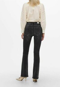 Black flared jeans made of denim with a smooth texture. Features two back pockets and stitched detailing. Worn with a light-colored blouse.