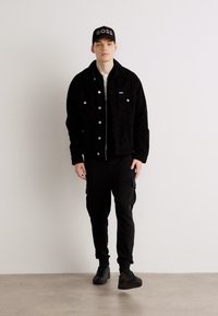 Schwarze Cordjacke mit vorderen Taschen, über einem schwarzen Hemd getragen. Cargo-Hosen und schwarze Schuhe runden das Outfit ab. Eine schwarze Mütze mit dem Text "BOSS".