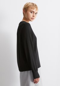 Μαύρο πλεκτό πουλόβερ με μακριά μανίκια, στρογγυλή λαιμόκοψη και ριβέ επωμίδες. Έχει χαλαρή εφαρμογή και φοριέται με ανοιχτό γκρι παντελόνι με παραλληλόγραμμες ρίγες.