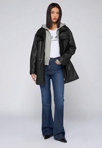 Czarny, oversize'owy, skórzany płaszcz na szarej bluzie z kapturem i białym T-shircie, zestawiony z dzwonowatymi niebieskimi dżinsami i czarnymi, szpiczastymi butami.