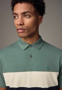 Teal värvi polo särk, millel on kolmes toonis disain heledad hallid ja tumesinised triibud, nööbid kaeluses ja rinnal väike broderitud logo.