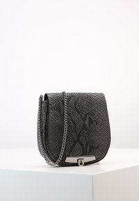 Mustan tekonahkaisen käärmeenahkakuvioidun olkalaukun kaareva siluetti, hopeaketjuside ja metallinen lukon yksityiskohta. Pinta on teksturoitu ja lisää syvyyttä.