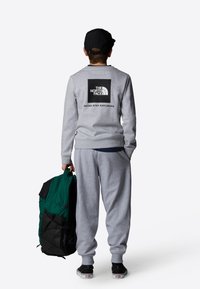 Grå hettegenser med svart logo på ryggen, grå joggebukser og en grønn og svart ryggsekk. Personen har på seg en svart caps og svarte sko.