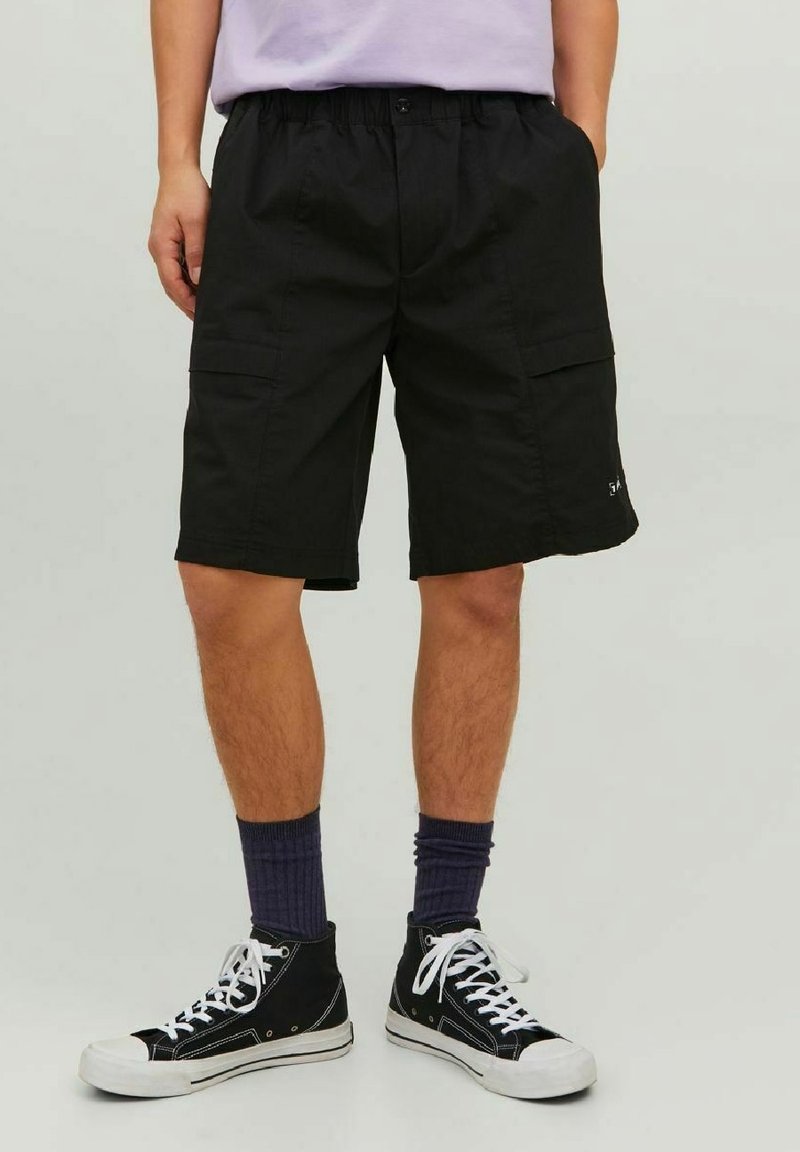 Personne portant un short cargo noir, des chaussettes hautes foncées et des baskets montantes noires avec des lacets blancs, debout devant un fond uni.