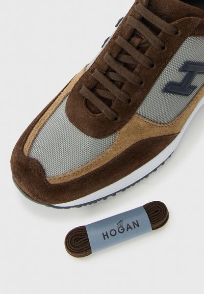 Brown and beige suede sneaker with gray mesh, brown laces, and extra rolled brown laces labeled "Hogan" on white background.