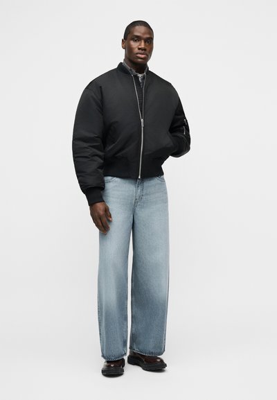 Άνδρας που φοράει μαύρο μπουφάν bomber, ανοιχτό μπλε τζιν με φαρδιά μπατζάκια και σκούρα καφέ παπούτσια, στέκεται μπροστά σε απλό ανοιχτό φόντο.