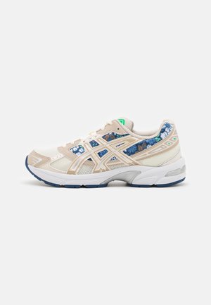 Chaussure de course beige clair et blanche avec motifs floraux bleus et bandes de logo beiges, vue de l'intérieur sur un fond uni.