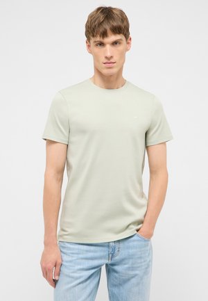 Junger Mann mit kurzen braunen Haaren, der ein hellbeiges kurzärmeliges T-Shirt und hellblaue Jeans trägt, steht mit einer Hand in der Tasche.