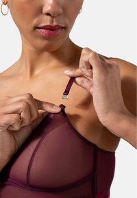 Rinna läpinäkyvä bralette, jossa on säädettävä hihna ja metallinen koukku. Lähikuva kädestä, joka tarttuu hihnaan, korostaen sen rakennetta ja istuvuutta.