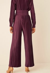 Pantalons larges plissés prune avec une taille ajustée. Le tissu a une texture lisse et des rayures verticales, complétant une silhouette haute couture.
