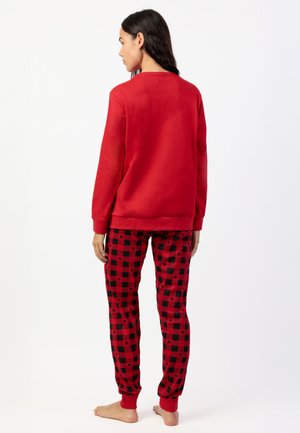 Donna in piedi a piedi nudi di spalle, indossa una felpa rossa e pantaloni del pigiama a quadri rosso e nero con piccoli motivi neri.