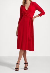 Robe portefeuille rouge avec un décolleté en V profond, manches longues et ceinture nouée ; fabriquée en tissu lisse, longueur au genou, associée à des brides noires autour de la cheville.