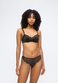 Femme aux cheveux bouclés portant un soutien-gorge en dentelle noire à fines bretelles et une culotte assortie en dentelle transparente, debout devant un fond clair uni.