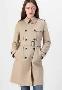 Beige trenchcoat met een dubbelknopens ontwerp, grote zwarte knopen, een ceintuur in de taille en schouderepauletten; gladde textuur.
