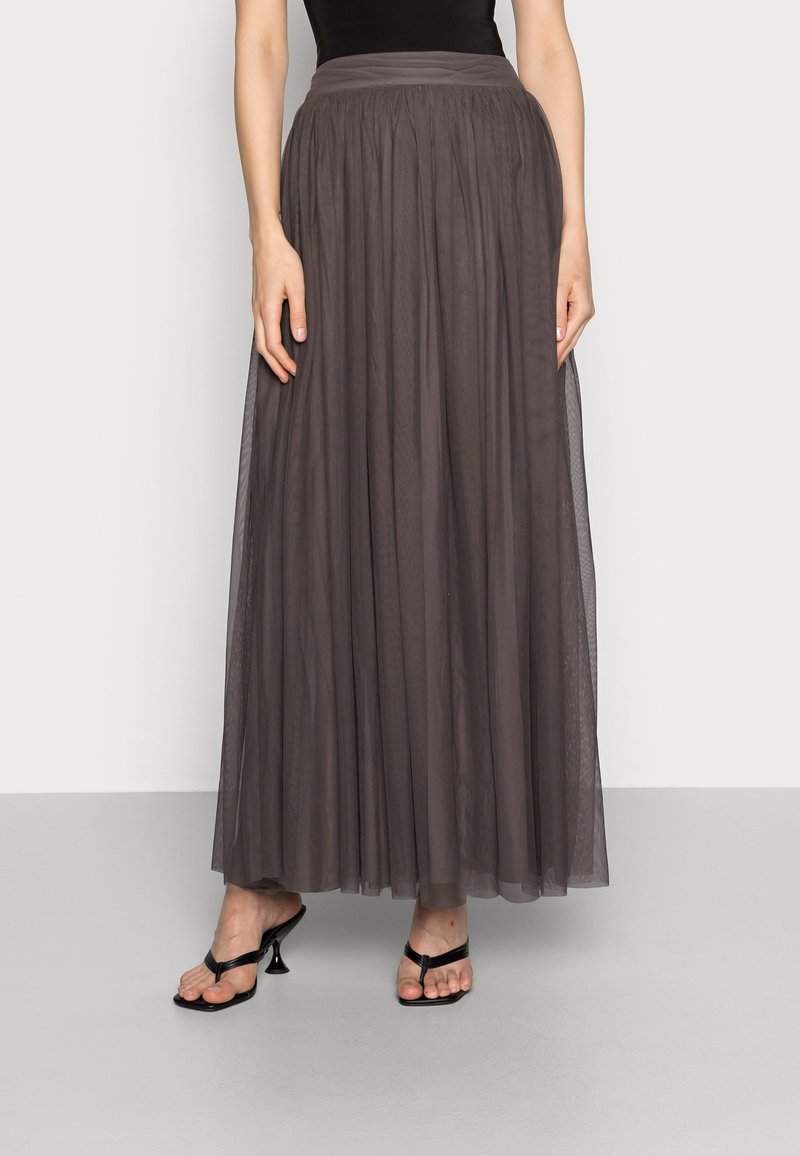 Femme portant une longue jupe transparente gris foncé avec une taille ajustée et des sandales noires à talons ouvertes à l’avant, debout devant un fond uni.
