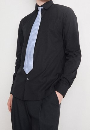 Personne portant une chemise noire et un pantalon avec une cravate à rayures bleu clair, debout avec une main dans la poche sur un fond uni.