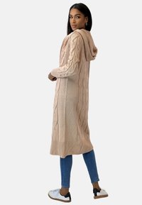 Lång kofta i kabelstickat ljusrosa med huva. Ribbstickade muddar och fåll. Bärs över tajta jeans och avslappnade sneakers, vilket visar lager på lager-textur.