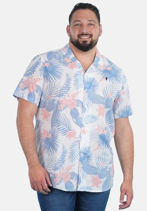 Homme aux cheveux courts et foncés et à la barbe, portant une chemise à manches courtes boutonnée avec des motifs floraux et de feuilles bleus et roses.
