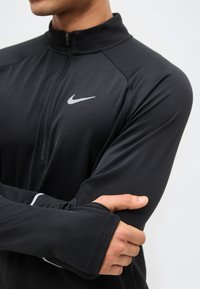 Άνδρας που φοράει μαύρο αθλητικό τοπ μακρυμάνικο της Nike με τρύπες για τον αντίχειρα, σταυρωμένα τα χέρια και ορατό το λογότυπο της Nike στο στήθος.