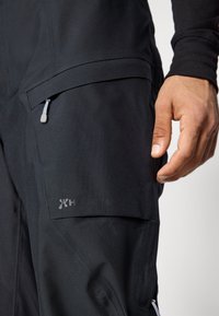 Crne vodootporne hlače glatke teksture, sa bočnim džepovima i patentnim zatvaračem. Na bedro je tiskani brendirani logo.