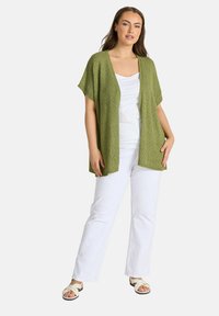 Mujer de pie con camiseta sin mangas blanca, pantalones blancos, sandalias blancas y un cárdigan verde suelto abierto al frente con mangas cortas.