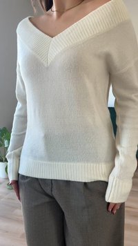 Off-white knitted sweater with a wide ribbed V-neck and hem, featuring long sleeves and a soft texture, paired with gray pleated trousers.