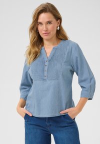 Blouse en chambray bleu clair avec un design plissé, encolure en V, manches trois-quarts et cinq boutons métalliques à l'avant. Tissu doux et texturé.