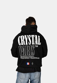 Felpa nera con cappuccio caratterizzata da grande testo bianco "CRYSTAL PARIS™" sulla schiena, con una piccola bandiera francese e logo a diamante in basso.