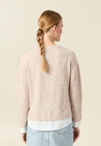 Maglione a maglia rosa chiaro con scollatura rotonda, orlo a coste e spalle scese. Indossato sopra una camicia bianca con colletto. Texture liscia.